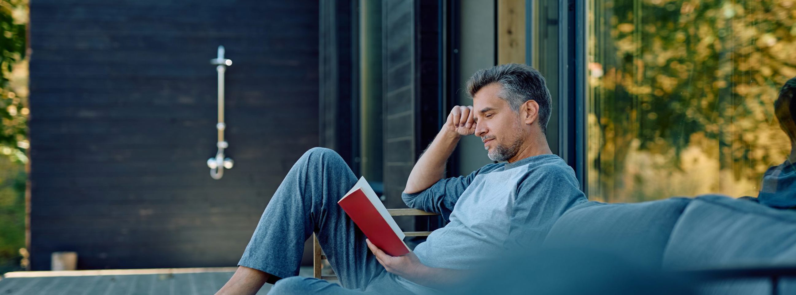 En mand sidder på terrassen i sit sommerhus og læser en bog.