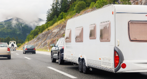 Sort personbil med campingvogn spændt bagpå kører på motorvej i et bjerglandskab.