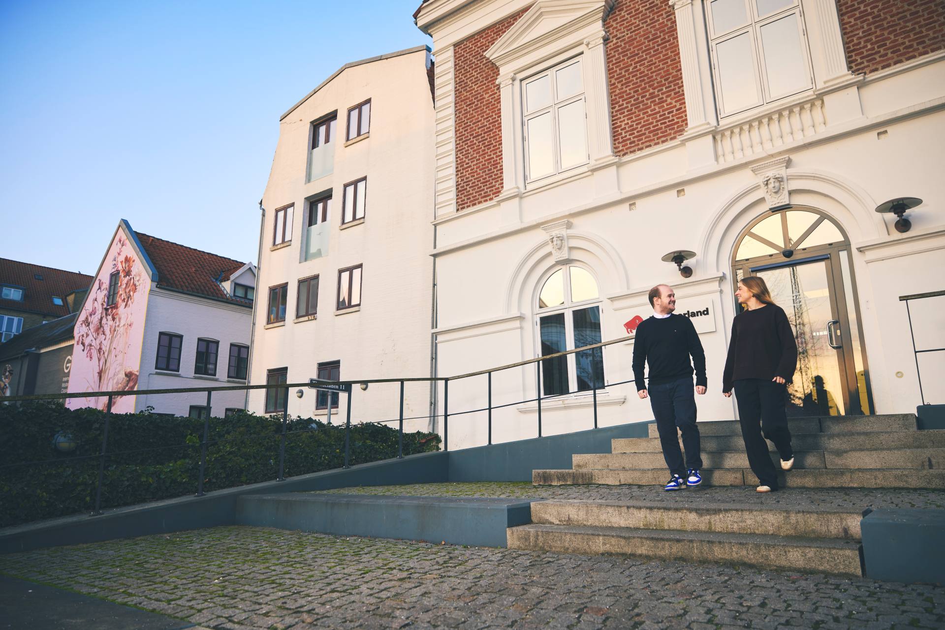 To unge mennesker går udenfor en forsikringsbygning og smiler til hinanden