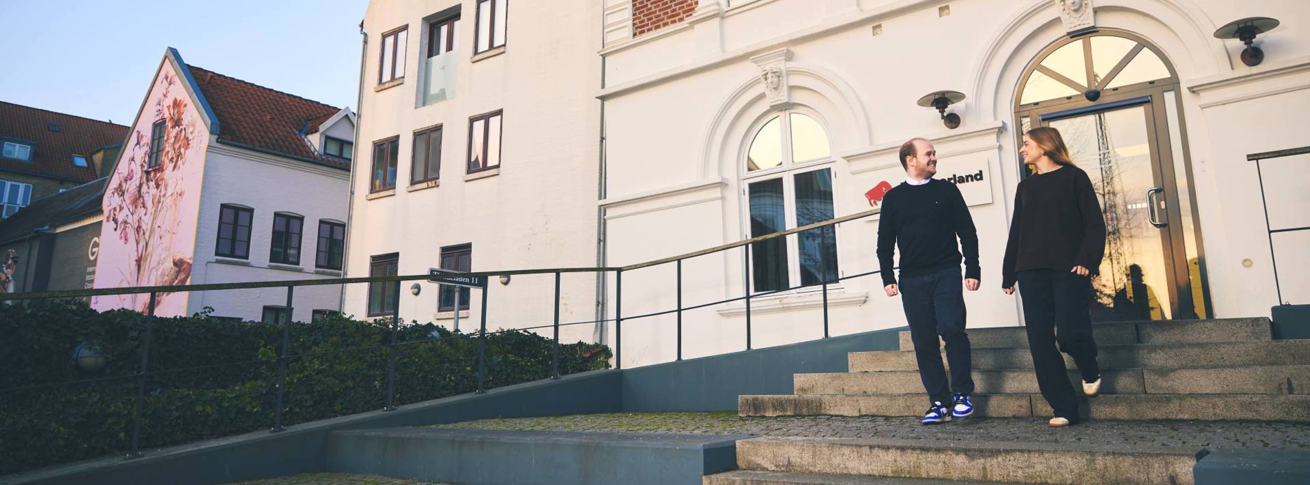 To unge mennesker går udenfor en forsikringsbygning og smiler til hinanden