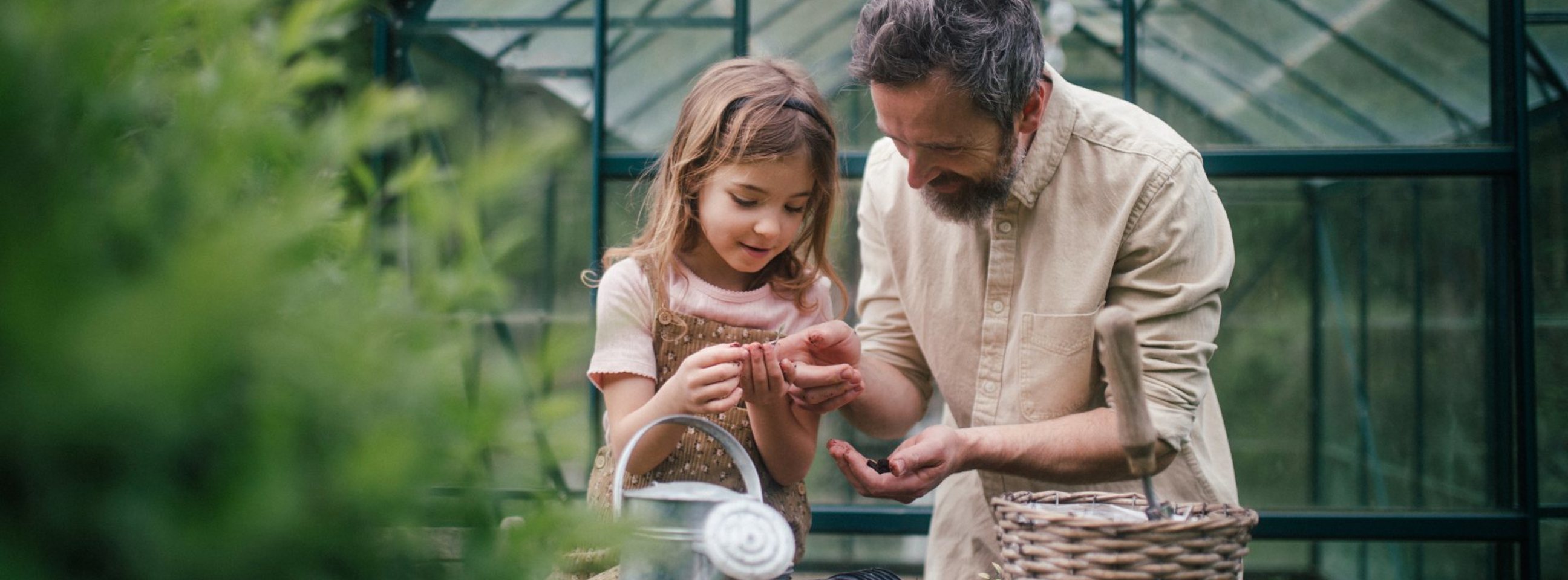 En far og en datter står foran et drivhus. På et bord står en vandkande og forskelligt andet haveudstyr.